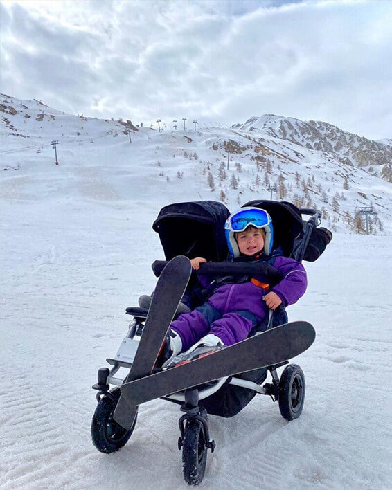 child dressed in ski suit with both skis on sitting in duet™ four wheeled double pram at skiing field - Mountain Buggy life without limit